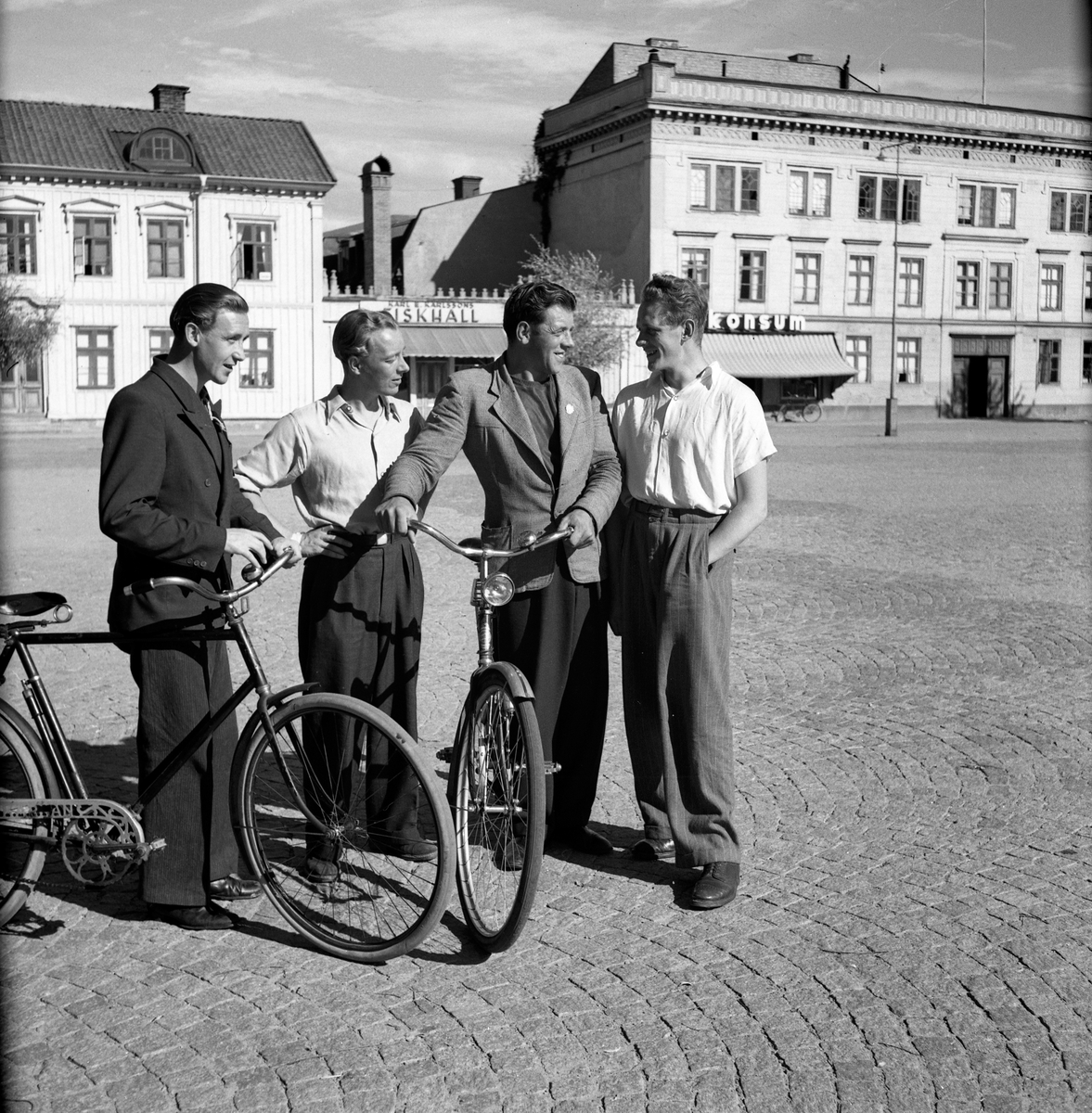 På Östra torget i Jönköping står fyra män med två cyklar. De två i mitten är från vänster Gösta Fröding och Olle Lindberg. I bakgrunden syns Amerikahuset.