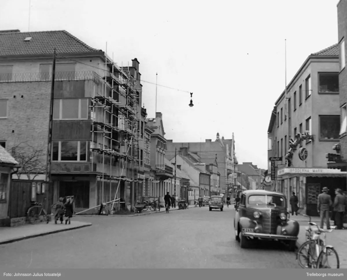 Algatan. Korsningen Algatan-Valldammsgatan. Vy mot ost. Till vänster, i kvarteret Kvadraten, byggs Schlyterska huset. Schlyters möbler flyttade in på bottenvåningen. Sedan kom under 60-talet radio- och TV-företaget B O Radio, sedermera Daler. Sedan övertog Jarl Svensson fastigheten och Modehuset Signum flyttade in. Till höger om Schlyterska huset ligger Apoteket Svea. Till höger i bild, i kvarteret Oden, ligger Biografen Röda Kvarn.