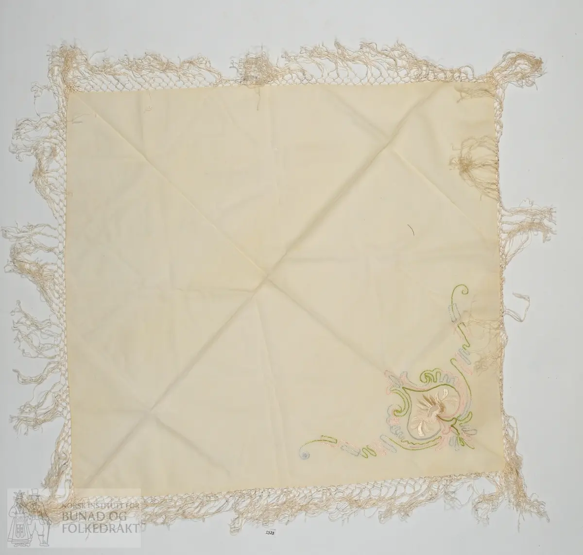Mjukturkle i kvit ubleika ull med taburerig i blått, rosa og grønt, samt blomst i kvitplattsaum. Frynser langs kanten, truleg av viskose.
Mål 70x70cm