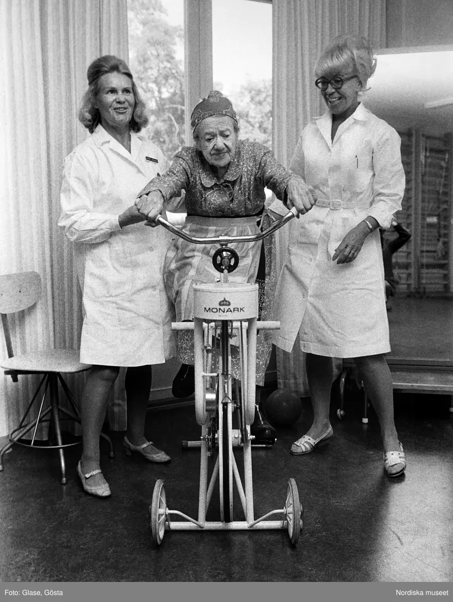 En gammal kvinna på motionscykel av märket Monark flankerad av vårdpersonal i vita rockar.