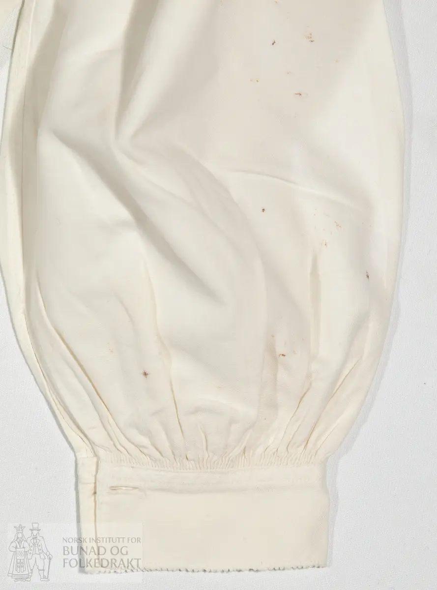 Skjorte i kvit bomull med musekantar "melegg". Maskinsydd og handsydd. Høg krage.

Mål:
Vidde 120cm. 
Rygglendeidde 46 cm. 
Høgde under arm 14 cm. 
Firkant u. arm 7cm x 8cm. 
Armlinning 4 cm. 
Krage 7, 5.
