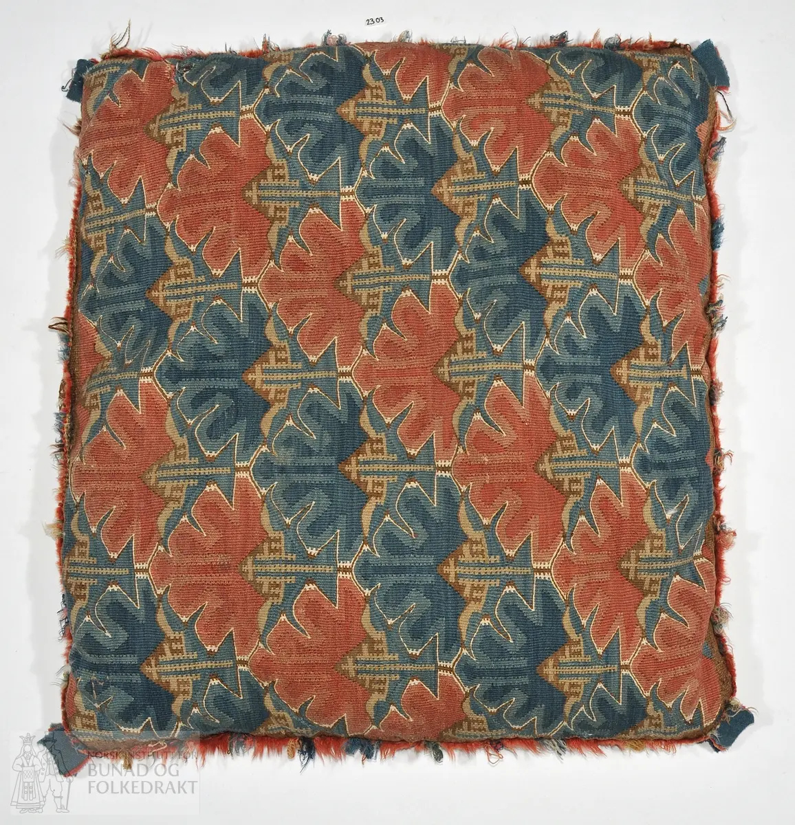 Pute av vevd ullsstoff etter billedvevprinsippet i ulike farger. Skinn på baksida. Fora med fjør og dun. 60 x 60