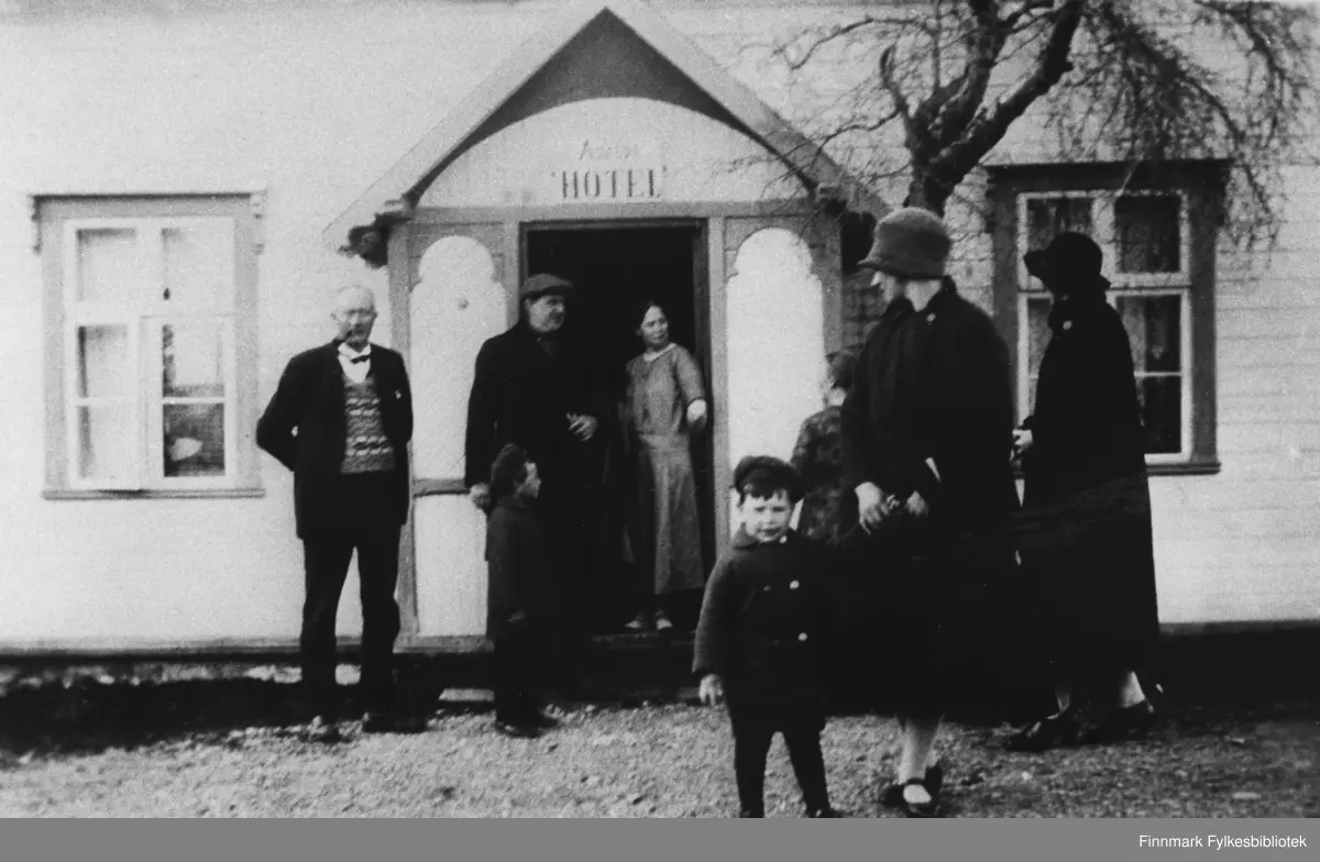 Astrups hotell på Langnes i Tana. Del av en serie bilder fra Langnes. Fotografert av Knut Grimm Fixdal. 

Thormod Holti skriver følgende om dette bildet i nettsamfunnet "Gamle bilder fra Tana": 

Oddmund Jessen forteller følgende om bildets innhold: 

I døråpningen står hotelleierJohanne (Lulla) Astrup (f.09.01.1879, d. 11.04.1941) som er datter til Christian Garmann Astrup og søster til Marie Amalie Astrup, mor til søstrene Øwre) 

Til venstre for Johanne (Lulla) Astrup står overingeniør i vegvesenet i Finnmark ,Jacob Natvig Matzow (f.22.06.1890 i Trondheim). Matzow hadde hytte på Langnes og bodde i Vadsø. 

Jakob Natvig Matzov var bygningsingeniør og tidligere broingeniør i U.S.A. Deltok under 1. verdenskrig med de amerikanske ingeniørtropper i Frankrike som mesteringeniør, senior grad. Avdelingsingeniør og veisjef for Statens veivesen i Finnmark. Etter 2. verdenskrig overingeniør for Finnmarks gjenreisningssjef i Vadsø. Han var også veisjef i Akershus fylke. Matzov mottok Kongens fortjenstmedalje i gull. (se her: http://www.stormo.de/AnnenVeginfo/VegsjeferFinnmarkPDF.pdf)

Mannen til venstre er overingeniør Knut Grimm Fixdal  (se også GK-FV.0000733) som også jobbet i Veivesenet rett under Veidirektøren og bodde i Vadsø. De to guttene er Fixdals sønner, hans kone Aagot Charlotte, født Øwre, holder den ene gutten i hånden. Knut Grimm Fixdal, født 16.09.1887 i Vestnes. Sønn av forsorgssjef Nils A. Fixdal og Serianna Stokkeland. Eks. T.T.L. 1911. Ekstraingeniør Vegdirektoratet 1911, ekstraingeniør i Bratsberg amt 1911-13, assisterende ingeniør i Finmarkens amt 1913-17, teknisk konsulent i Haugesund 1917-20, avdelingsingeniør i Finnmark fylke 1920, overingeniør og sjef for vegvesenet i Finnmark fylke 1920-37, overingeniør (vegsjef) i Telemark fylke 1937-57. I 1945 midlertidig fylkesmann i Telemark fylke. Medlem av Vadsø bystyre, formann N.I.F. Finnmark avd. Mottok Kongens fortjenestemedalje i gull. 

Den andre kvinnen er Gudrun Johanne Matzow, født Øwre, gift med Jakob Natvig Matzow. 

Gudrun Johanne og Aagot Charlotte var døtrene til Marie Amalie Astrup (født 20.11.1865, død 14.11.1923) og Nils Øwre (Nils Øvre, kjøpmann født 04.04.1861 i Lebesby, død 16.01.1937,  fiskeforretning i Mehamn, Gamvik fra 1905, senere landhandel i Tana 1888-1918, periodevis lensmann i Karasjok og rettstolk i lappisk og kvænsk. Øwre hadde flere kommunale tillitsverv ). 
(For Øwre se her: http://runeberg.org/merkbio/0936.html) 
: 
(Om Fixdal, se http://www.stormo.de/Veghistorie/Organisasjon/Organisering%201923-1930.pdf)

Dette bildet har brukt i flere sammenhenger, sist i boka "Sentrum ved Flodmunningen" skrevet av Thormod Holti og Dag F. Simonsen på side 83.

