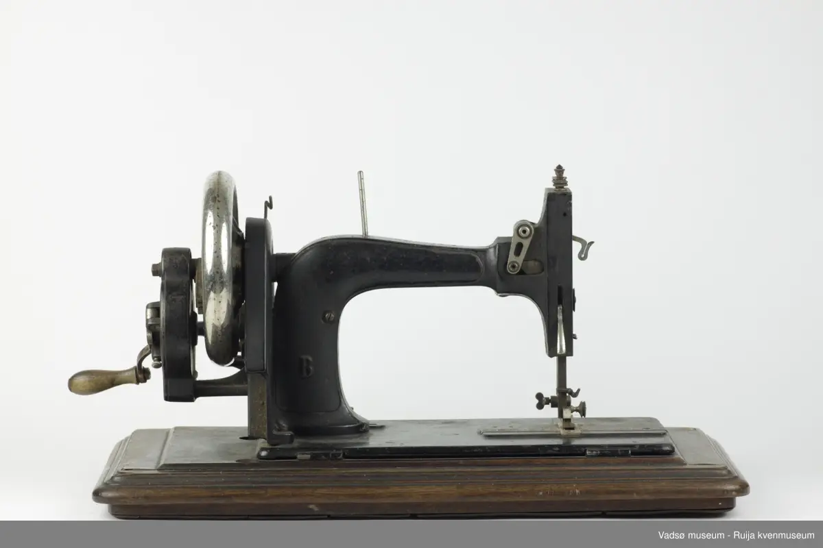 Hånddrevet symaskin av typen Model B, produsert i 1896 av G.M. Pfaff Kaiserslautern, Tyskland. Maskinen er hengslet fast i en profilert sokkel i treverk. Inni sokkelen finnes et rom beregnet til diverse symateriell. Rommet nås gjennom å løfte maskinen fra brukersiden. I sokkelen, parallelt med maskinens lengderetning, er det innfelt en målestokk.