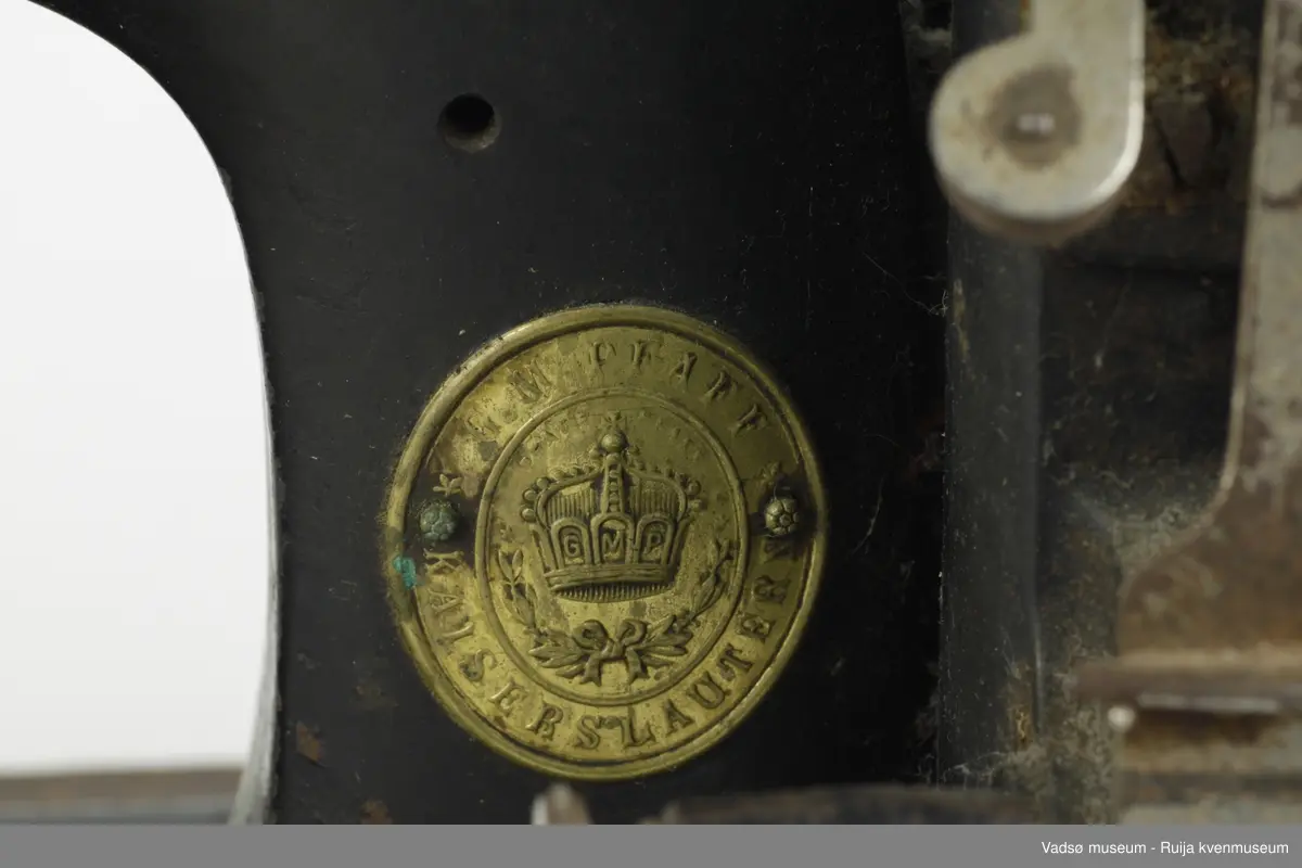 Hånddrevet symaskin av typen Model B, produsert i 1896 av G.M. Pfaff Kaiserslautern, Tyskland. Maskinen er hengslet fast i en profilert sokkel i treverk. Inni sokkelen finnes et rom beregnet til diverse symateriell. Rommet nås gjennom å løfte maskinen fra brukersiden. I sokkelen, parallelt med maskinens lengderetning, er det innfelt en målestokk.