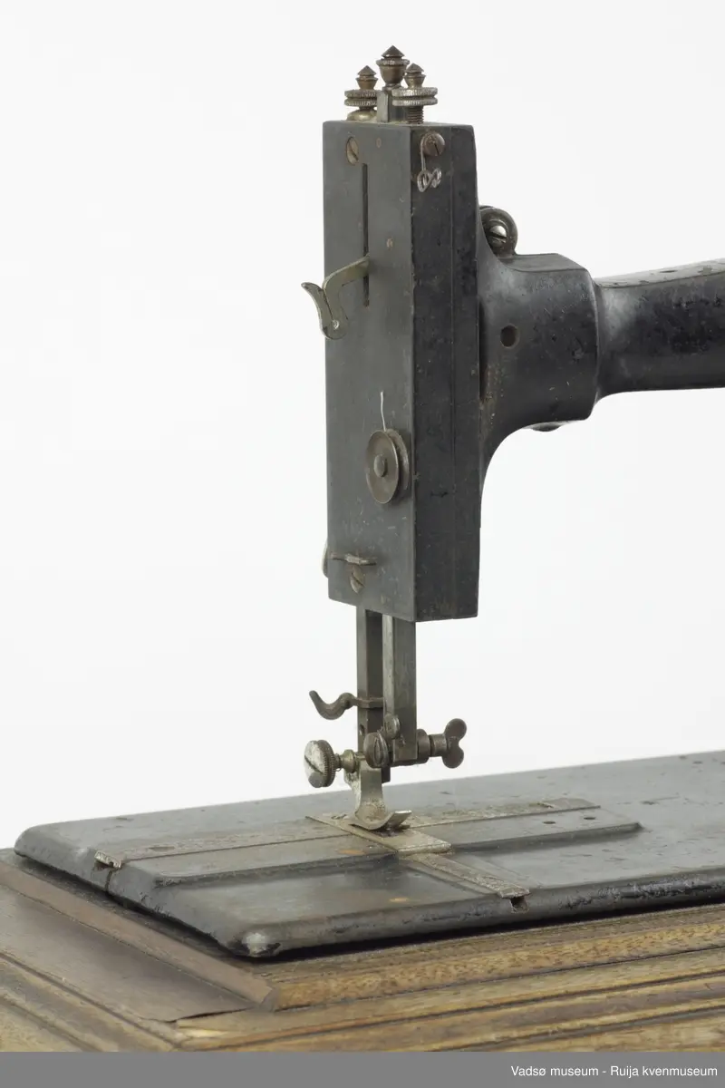 Hånddrevet symaskin av typen Model B, produsert i 1896 av G.M. Pfaff Kaiserslautern, Tyskland. Maskinen er hengslet fast i en profilert sokkel i treverk. Inni sokkelen finnes et rom beregnet til diverse symateriell. Rommet nås gjennom å løfte maskinen fra brukersiden. I sokkelen, parallelt med maskinens lengderetning, er det innfelt en målestokk.