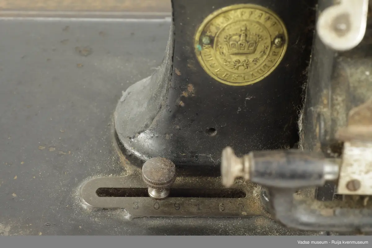 Hånddrevet symaskin av typen Model B, produsert i 1896 av G.M. Pfaff Kaiserslautern, Tyskland. Maskinen er hengslet fast i en profilert sokkel i treverk. Inni sokkelen finnes et rom beregnet til diverse symateriell. Rommet nås gjennom å løfte maskinen fra brukersiden. I sokkelen, parallelt med maskinens lengderetning, er det innfelt en målestokk.