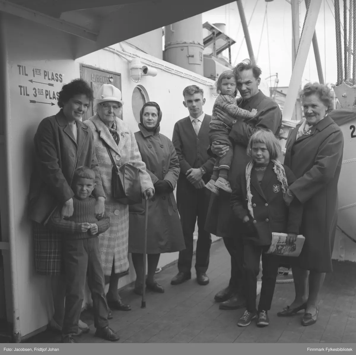 En familie poserer for fotografen ombord på et skip,  tatt i august 1964. Skipet har tredekk og "naglede livbåter". På veggen bak familien står det anvisninger til 1.og 3.plass med piler og tekst.  Fra venstre: Herbjørg Jacobsen med sønnen Tom Jacobsen rett foran seg. Ved siden av står Olga Jacobsen med en stokk i sin venstre hånd og håndveska hengende foran. Aase Jacobsen har en ganske mørk kåpe, svarte hansker og tørkle på hodet. Åge Christian Jacobsen iført mørk bukse og jakke med lys skjorte og slips. Alf Edvard Jacobsen med datteren Eli på armen. Han er iført mørk frakk og bukse og Eli har mønstret strikkegenser på seg. En ukjent jente er iført mørk kåpe, mørke bukser og older i et blad. Damen lengst til høyre er Eli Tverrfjell, moren til Herbjørg. Dekket har tregulv og en livbåt ses helt til høyre på bildet. Master, tauverk og stag ses bak på bildet.