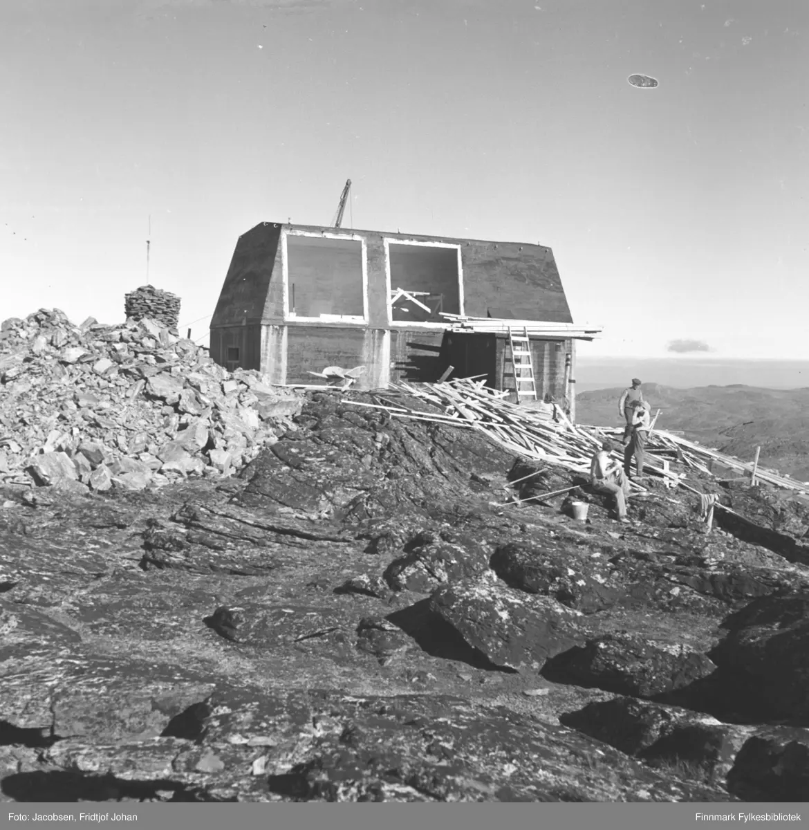 Arbeidskarer på fjellet Tyven i Hammerfest. Til venstre står varden. Forsvarets radiolinjestasjon er under bygging og en stabel med material ligger ved siden av det. Det er antakelig forskalningsmaterialene som er revet av etter betongen har tørket. De var der før Telegrafverket fikk sin i gang noen år senere. En haug med steiner ligger til venstre på bildet. Bygget står i dag i nærheten av TV-masta som sannsynligvis er under bygging.