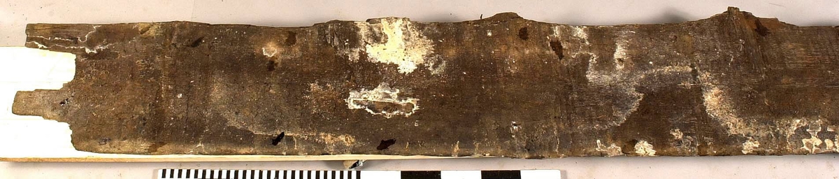 Däcksbräda.
Ena änden är skadad och partier saknas. I den andra änden börjar en genomgående spricka som löper utmed halva brädan. Skador och brott längs ena långsidan. Genomgående och ytliga sprickor.