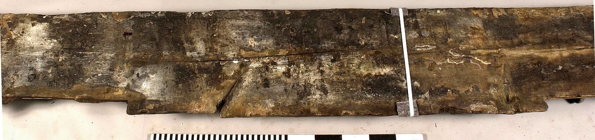 Däcksbräda.
Ena änden är skadad och partier saknas. I den andra änden börjar en genomgående spricka som löper utmed halva brädan. Skador och brott längs ena långsidan. Genomgående och ytliga sprickor.