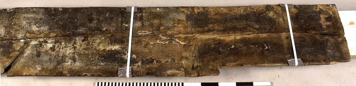 Däcksbräda.
Ena änden är skadad och partier saknas. I den andra änden börjar en genomgående spricka som löper utmed halva brädan. Skador och brott längs ena långsidan. Genomgående och ytliga sprickor.