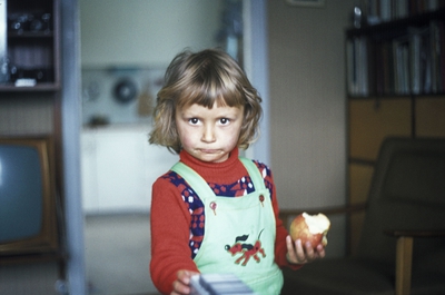 Lill Carmen Jensen. Hun har på seg en genser med mønster og bukse med seler. Buksen har et hundemotiv i fronten. Hun har et eple i hånden som det er tatt noen tygger av. Noe diffust kan man se en lenestol  og bak/over den en bokhylle med bøker i. Til venstre et TV-apparat og en hylle med noen pyntegjenstander over det.