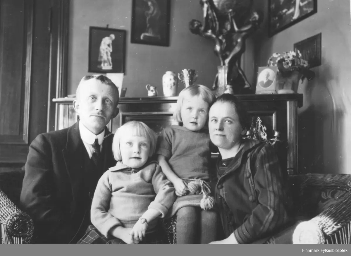 Fotografi av August Magnus Jacobsen fra Vardø og hustru Solveig Johannessen Jacobsen fra Bergen. På bilde er de sammen med sine døtre Gerd og Liv Vibe Jacobsen. De sitter i kurvstoler foran en peis. Oppe på ovnen står blant annet noen fotografier, noen vaser, en blomst og en stor bronsestatue av flere mennesker. På veggen henger det flere bilder. Bak August Magnus er det en dør