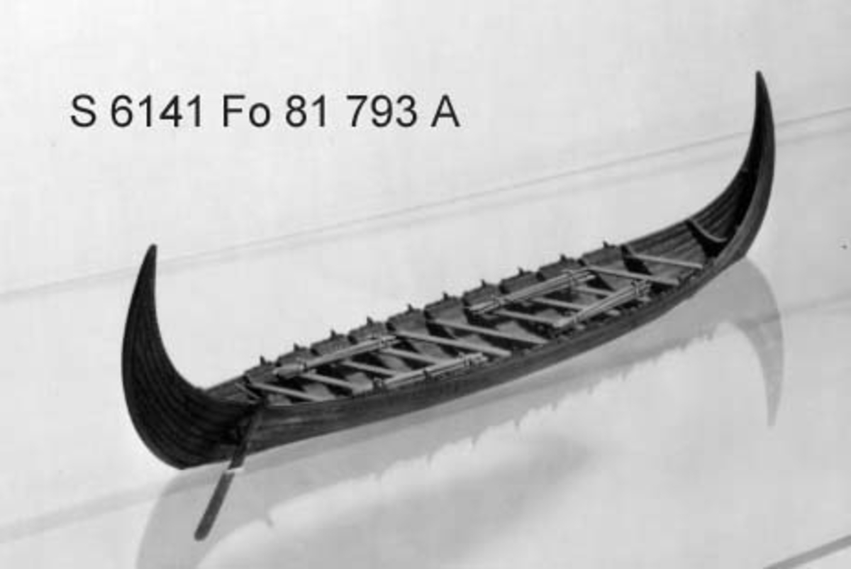 Fartygsmodell i oxelträ, rekonstruktion av "Stora Kvalsundsbåten, utgrävd 1920, daterad till 400-700 e.Kr.. Skrov utfört i två halvor med limfog genom köl och stävar. Bordläggningen skuren på såväl in- som utsidan. Klamparna på borden pålimmade. Håarna av lövträkvistar, med självvuxen knagge, fastbundna med svartoxiderad suturtråd. Skrovet betsat i tjärfärg.