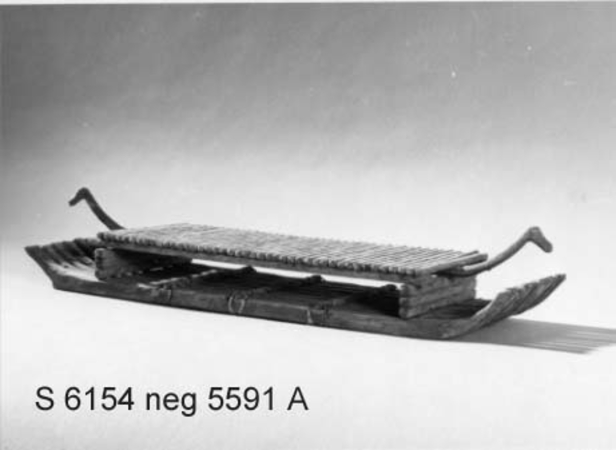 Modell av flotte, rekonstruktion av "Bredarörtypen", skeppsbild från Bredarör invid Kivik, Skåne. Flytkropp av 9 raka, i ändarna svagt uppböjda stockar, sammanhållna av 3 tvärbalkar och två plattformsstöttor. Plattformen består av tvärgående ribbor uppburna av tre längsgående spärrar varav den mittersta, utskjutande, avslutas med djurhuvud. Skala: 1/60 (Jfr. S.5892 = skala 1/50).