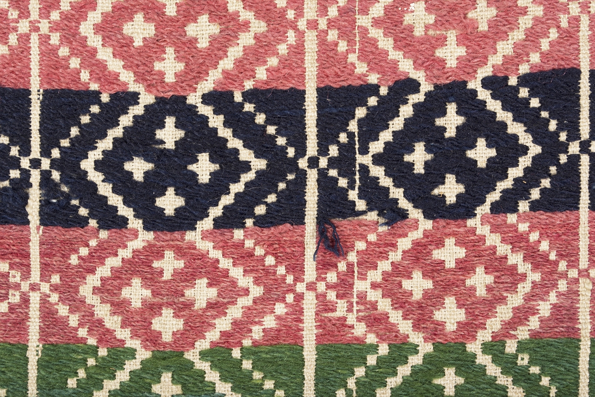 Randigt täcke vävt i opphämta. Täcket är randigt på bredden i inslagsfärgerna rosa och mörkblått, varje rand är 43 mm bred. Ränderna på längden löper som sicksack-bårder med fyra kors i grupp inom varje rombformation. Runt om löper en grön bård i samma mönster som täcket i övrigt. Mönsterinslag i ullgarn på tuskaftsbotten i oblekt lingarn. Smal fåll i kortsidorna, stadkanter i långsidorna. Täcket är ihopsytt på mitten av två delar.Varp i oblekt 1-trådigt z-spunnet lingarn, 13,5 trådar/cm.Botteninslag i oblekt 1-trådigt z-spunnet lingarn, 10,5 trådar/cm.Mönsterinslag i 2-trådigt z-tvinnat ullgarn.Märkt på baksidan med en påsydd tyglapp med texten: "N° 154 a Rönnebergs".Täcket har också varit märkt med en fastnästad brun pappersetikett (nu förvarad i reg.pärm) från Svenska Hemslöjdsföreningarnas Riksförbund, med tryckt och maskinskriven text: "Föreningens namn: Malmöhus läns Hemslöjdsförening Malmö. Föremålets art: Gammal täcke Rönneb. h:d. Föremålets storlek: 120 + 165 Teknik: opphämta Teknikens ortsbenämning: Skälblad Material: Linne & ullgarn. Försäljningspris: -  Kopia, bearbetning eller komposition: - Utfört av:  - ".