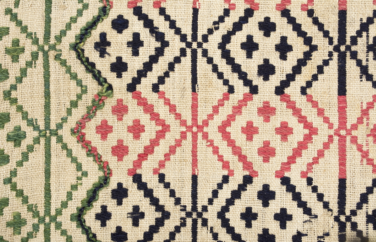 Randigt täcke vävt i opphämta. Täcket är randigt på bredden i inslagsfärgerna rosa och mörkblått, varje rand är 43 mm bred. Ränderna på längden löper som sicksack-bårder med fyra kors i grupp inom varje rombformation. Runt om löper en grön bård i samma mönster som täcket i övrigt. Mönsterinslag i ullgarn på tuskaftsbotten i oblekt lingarn. Smal fåll i kortsidorna, stadkanter i långsidorna. Täcket är ihopsytt på mitten av två delar.Varp i oblekt 1-trådigt z-spunnet lingarn, 13,5 trådar/cm.Botteninslag i oblekt 1-trådigt z-spunnet lingarn, 10,5 trådar/cm.Mönsterinslag i 2-trådigt z-tvinnat ullgarn.Märkt på baksidan med en påsydd tyglapp med texten: "N° 154 a Rönnebergs".Täcket har också varit märkt med en fastnästad brun pappersetikett (nu förvarad i reg.pärm) från Svenska Hemslöjdsföreningarnas Riksförbund, med tryckt och maskinskriven text: "Föreningens namn: Malmöhus läns Hemslöjdsförening Malmö. Föremålets art: Gammal täcke Rönneb. h:d. Föremålets storlek: 120 + 165 Teknik: opphämta Teknikens ortsbenämning: Skälblad Material: Linne & ullgarn. Försäljningspris: -  Kopia, bearbetning eller komposition: - Utfört av:  - ".