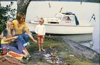 Albin 21 förtöjd i en vik. I förgrunden grillar en familj korv över öppen eld. Marknadsföringsbild.