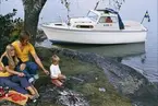Albin 21 förtöjd i en vik. I förgrunden grillar en familj över öppen eld. Marknadsföringsbild.