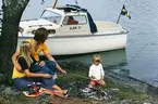 Albin 21 förtöjd i en vik. I förgrunden grillar en familj över öppen eld. Marknadsföringsbild.