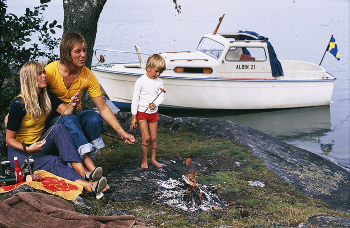 Albin 21 förtöjd i en vik. I förgrunden grillar en familj över öppen eld. Marknadsföringsbild.