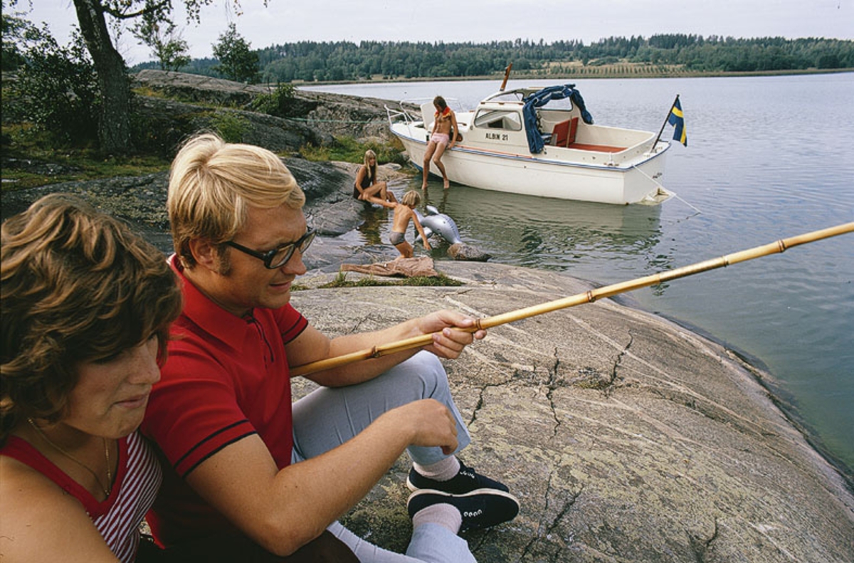 Albin 21 förtöjd i en vik. I förgrunden ett par med metspö. Marknadsföringsbild.