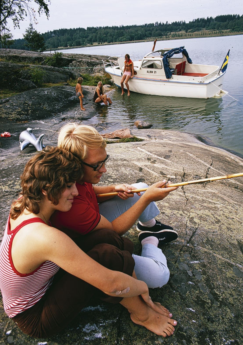Albin 21 förtöjd i en vik. I förgrunden ett par med metspö. Marknadsföringsbild.