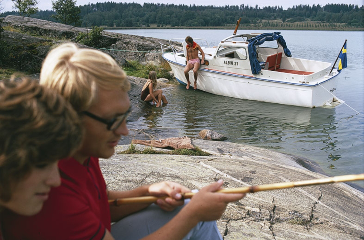 Albin 21 förtöjd i en vik. I förgrunden ett par med metspö. Marknadsföringsbild.