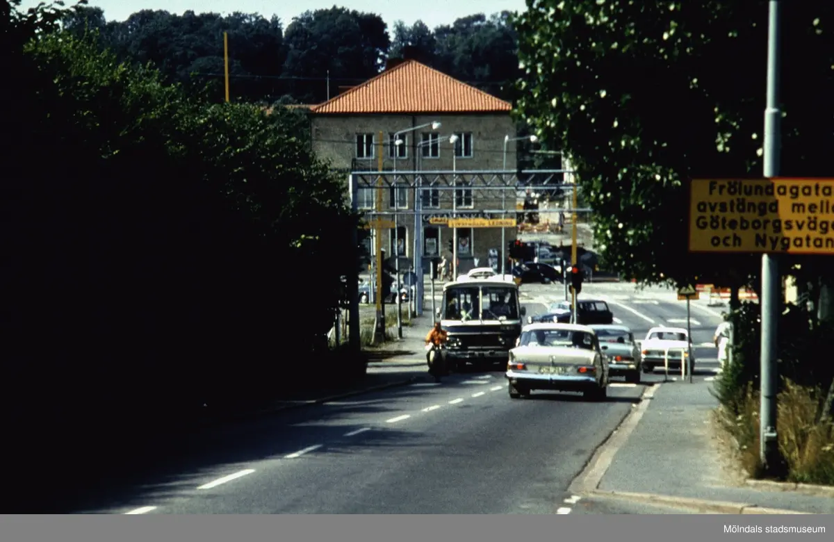 Vy från Kvarnbygatan mot uppresta järnvägsbommar där en buss och bilar passerar, 1970-tal. I bakgrunden ses Hantverkshuset (Kungsbackavägen 2/Frölundagatan 1) i Mölndals Bro.