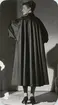 Nordiska Kompaniet. Kvinna i mörk kappa med mjuka veck i ryggen och raglanskurna vida ärmar. Text på baksidan: 