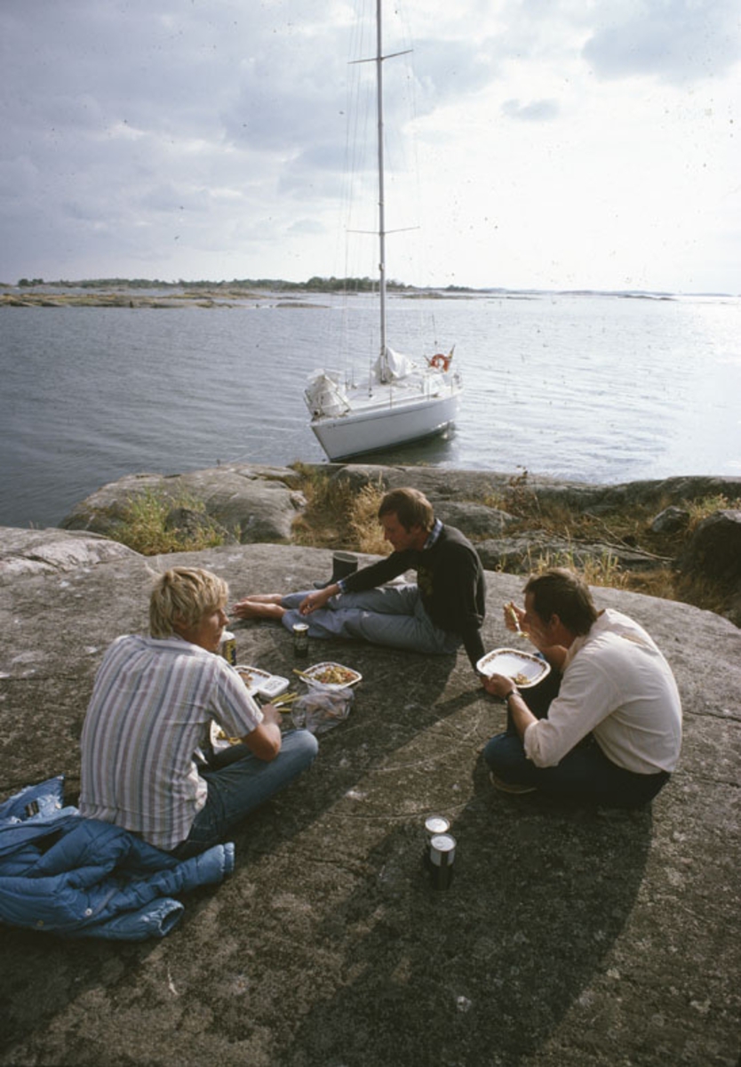 Tre män äter en måltid på en klippa. I bakgrunden en förtöjd Scampi 30.