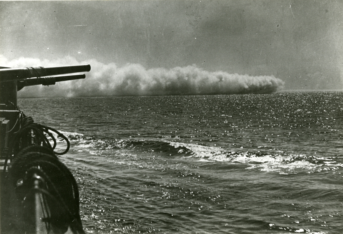 En svensk jagare lägger ut en dimridå för att skydda pansarskeppet i förgrunden.