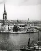 Vy över Stockholm från Mälarsidan, Skinnarviksbergen.
Vid Riddarhuskajen ligger passagerarångfartyget Tjust med (flera oidentifierade fartyg), Nya Södertalje (2 fr.v.), Nya Hållsviken (3 fr.v.), Sjöfröken (4 fr.v.).