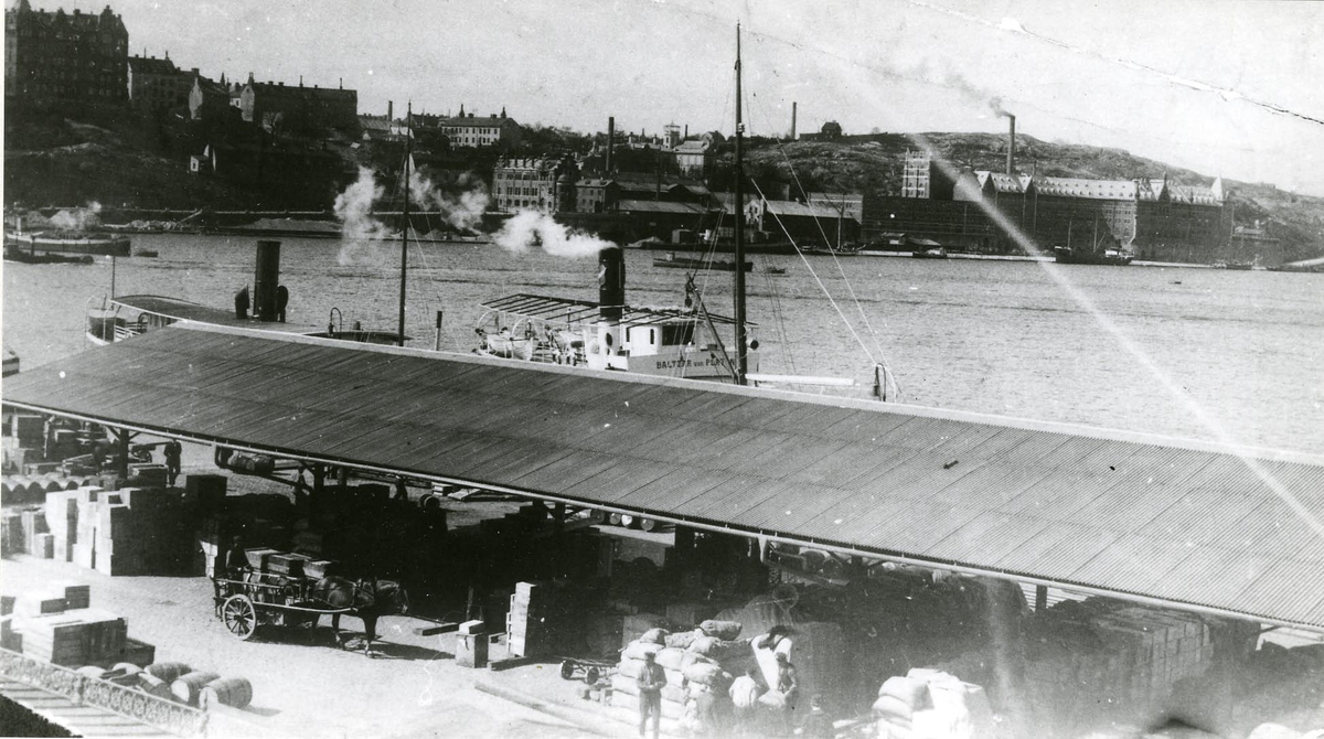 Riddarholmskajen. I förgrunden tak över varorna på kajen. Ångfartyget Baltzar von Platen vid kaj.
Söder Mälarstrand och Münchenbryggeriet i bakgrunden.