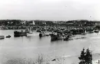 Livet vid brädgårdskajerna var livligt under utlastningssäsongen. Foto från ca 1930 taget från Öd och föreställande Kramfors östra brädgård. Hamnkontoret skymtar till höger och kanalens inlopp allra längst till höger i bild. I bakgrunden ligger Kramfors kapell. Tre virkeslastade ångare har ankrat upp framför brädgården, en av dem lastas från spetspråmar, ur vilka just hyvas några 