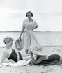 Kvinna i klänning, kort jacka och stråhatt i handen står på en strand. Två kvinnor i baddäkt ligger ned på stranden.