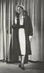 Modell i mörk kappa, vit klänning med brett skärp, vit hatt och vita handskar. Från J. Fath.