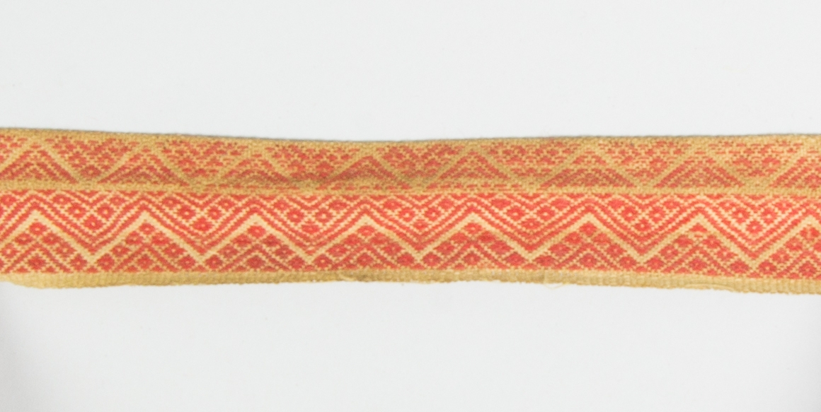 Kjolsäck till dräkt för kvinna från Mora socken, Dalarna. Modell med avskuret framstycke. Tillverkad av brunt skinn med dekor på framstycket och överstycket av remsor med stansat mönster av skinn, med underlägg av rött kläde som lyser genom, underläggssöm. På framstyckets band även en kant av grönt vadmal, nästan helt bortnött. Inlägg i sid- och bottensöm av två remsor tyg, rött kläde och grön vadmal. Mönstervävt band med plockat mönster i rött ullgarn på vit botten av lin eller bomull, gula kanter.