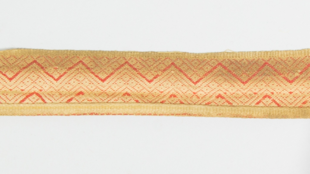 Kjolsäck till dräkt för kvinna från Mora socken, Dalarna. Modell med avskuret framstycke. Tillverkad av brunt skinn med dekor på framstycket och överstycket av remsor med stansat mönster av skinn, med underlägg av rött kläde som lyser genom, underläggssöm. På framstyckets band även en kant av grönt vadmal, nästan helt bortnött. Inlägg i sid- och bottensöm av två remsor tyg, rött kläde och grön vadmal. Mönstervävt band med plockat mönster i rött ullgarn på vit botten av lin eller bomull, gula kanter.