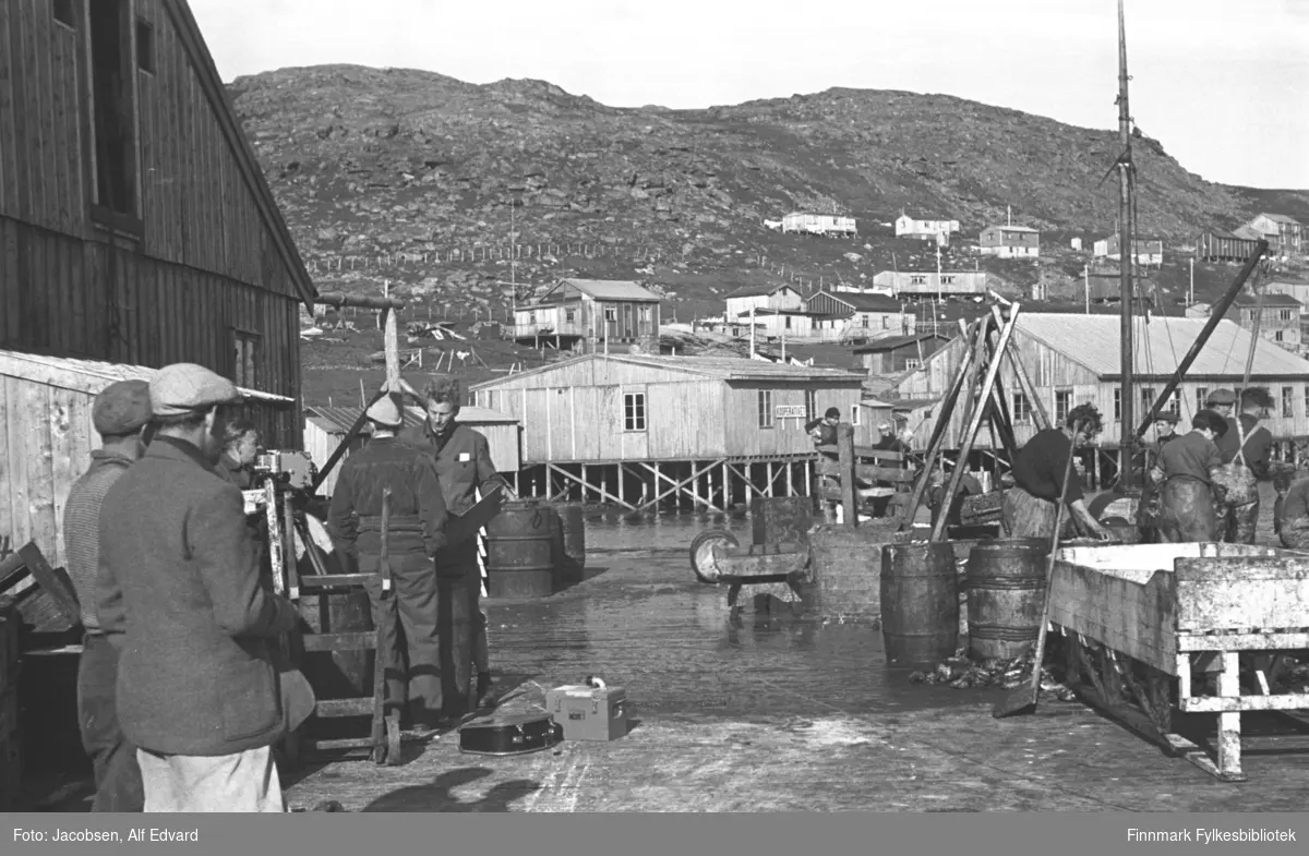 Bilde fra havneområdet i Havøysund. Bygget til venstre er Samvirklagets fiskebruk. Bygget har ganske mørkt, stående panel og en lasteluke oppe i røstet. Et lite tilbygg med skrått tak står på røstveggen og med to avlange vinduer på hver side. Flere menn er i arbeid. Mannen som er vendt mot kamera, ved veggen på fiskebruket er Åge Pedersen. De fleste har mørke klær, men mannen nærmste kamera har lys bukse og lys skyggelue. En stor og hvitmalt kasse ses nederst til høyre på bildet og to tønner av tre står ved siden av den.  Et par tønner/fat av metall står litt til høyre på bildet. Den umalte brakka, omtrent midt på bildet, som står oppå en trekai er Coopen brakka. Et mindre bygg står til venstre for det. Til høyre for Coopenbrakka står den litt større brakka som er Harald Olsens snekkerverksted. En mørkkledt mann lener seg mot det korte gjerdet foran Coopen. En mast med lastebom ses til høyre på bildet, sansynligvis en båt som ligger ved kaia. Noen hus og brakker er satt opp i lia ovenfor. Brakka som ses rett over taket på Coopen-brakka er huset til Gyda og Rolf Olsen. Huset til høyre for det er Seppola og litt opp og til høyre for Seppola, er huset til Alfred Jørgensen. Helt øverst, til høyre på bildet, er huset til Alfred Stabell som var det første permanente huset. Den litt lavere brakka til venstre for Stabell er brakka til Kollin Kristiansen. Fjellet og dalsiden har lite og ingen vegetasjon, men mye stein og et gjerde går på tvers av dalsiden.