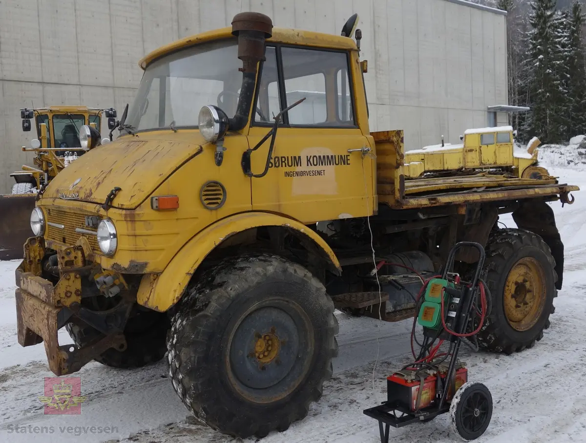 MB Unimog 406. Lastebil på 4 hjul, (to akslet), med akselavstand 240 cm. I hovedsak gul. 4-hjuls drift. Kjøretøyet har for fremdrift en 6-sylindret dieselmotor, antatt av typen M.B OM 352 X 11 fra Mercedes Daimler Benz. Motoren yter 84 Hk (61,76 KW) og har et slagvolum på 5700 kubikkcentimeter.
Schmidt standard frontmontert redskapsfeste. Antall seter er 2. Standard dekkdimensjon skal være 12,5-20 rundt om.