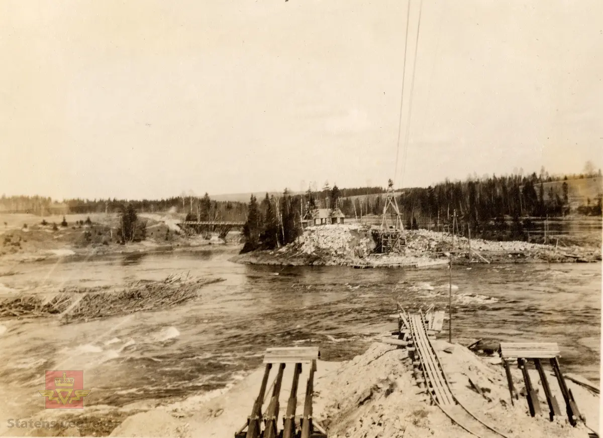 Bingsfoss bru.   I følge merking i album: "Utsikt fra søndre side, mot holmen m/tårn for taubanen. Denne blev montert våren 1924 over hovedløpet med sp.v.  215 m og bæreevne 3 t som enkellast - utstyrt med elektrisk drivkraft. Taubanen var til uvurderlig nytte på anleggets forskjellige trinn helt til hengebroen var montert.  I forgrunnen søndre kabelforankring færdig 1924. (140 m3 beton).  Fot. N.Sx."  I bakgrunnen bak holmen skimtes stillasbrua.