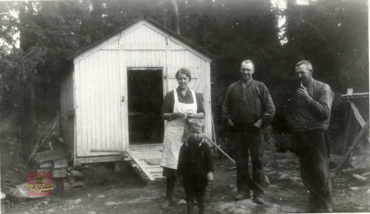 07.09.2015: "Midt på bildet står Oscar Andersen, født 1893, han jobbet som anleggsarbeider og skytebas  både ved byggingen av brua ved Rånåsfoss og Bingsfoss. Han var også med på et veganlegg på vegen fra Sandvika over Sollihøgda til Sundvollen. Ved siden av ham står hans kone Alma Andersen med hvitt forkle og strikketøy i hendene, og foran dem står adoptivsønnen Jørgen, omtrent 3 år gammel. Mannen til høyre med pipa er ukjent."  Informant til bildet er Bjørn Andersen.
Bildet er sannsynligvis fra Sollihøgda. Se også bilde NVM 00-F-03399.