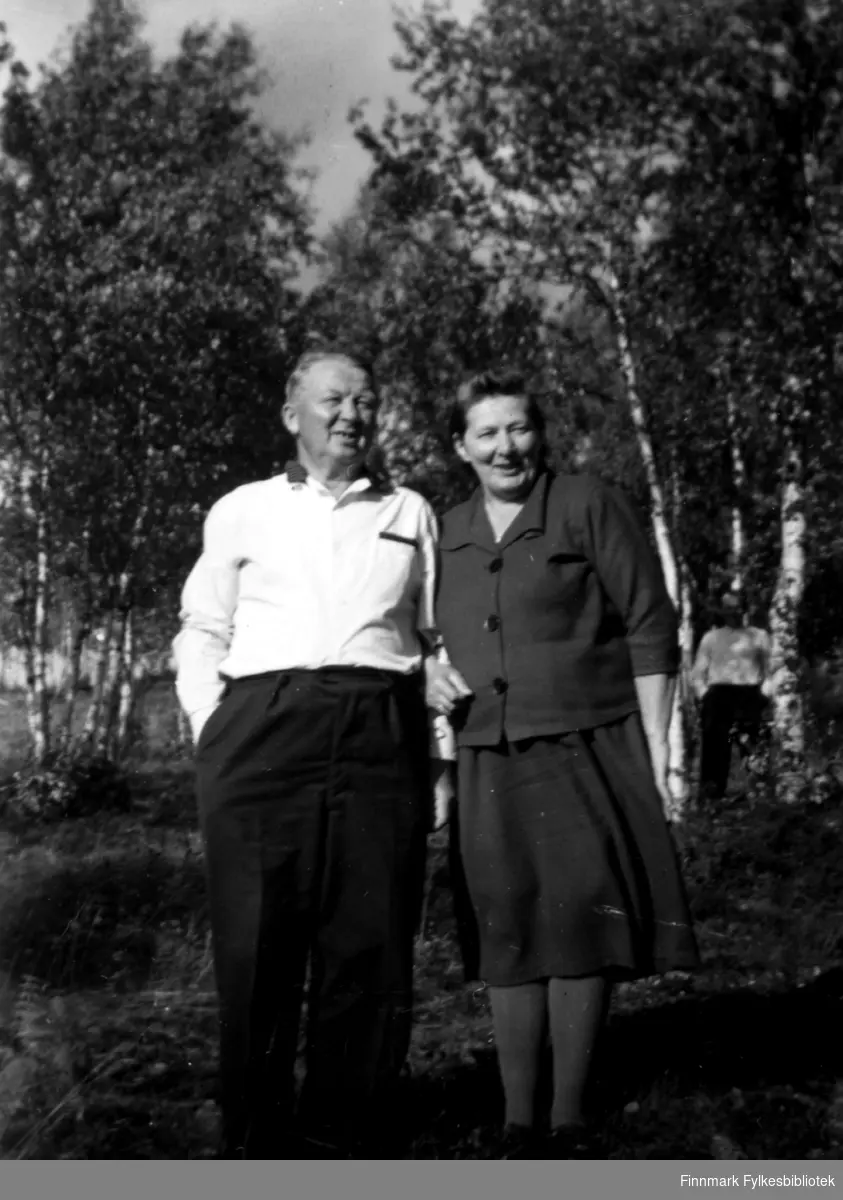 Fotografi av Johan og Henrika Sotkajærvi. Bildet er tatt ved en skog. Det er trær bak dem. Til høyre i bildet kan vi skimte en mann i en hvit skjorte og mørk bukse. Johan og Henrika holder hverandre rundt armen. Hun er kledt i en drakt. Johan er kledt i en hvit skjorte og mørk bukse