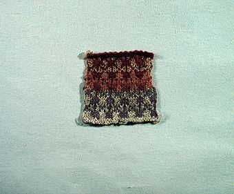 Stickprov i ullgarn. Slätstickad mönsterstickning i grått, blått och två nyanser brunt. Mönstret återfinns i Hermanna Stengårds "Gotländsk Sticksöm" nr. 25, sid. 45. Mönster från Tofta.