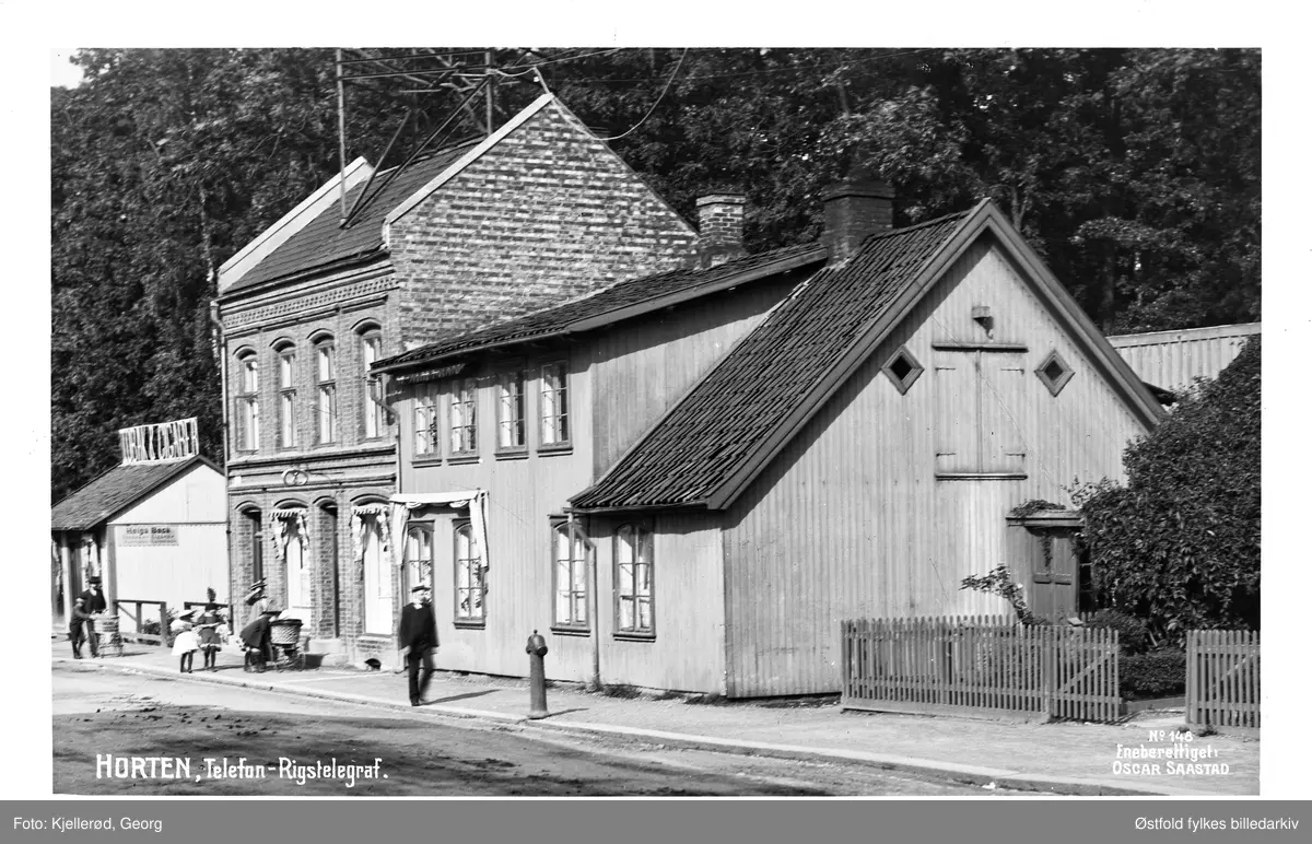 Horten med bygning for telefon og telegraf i Storgata. 

Helt til venstre: Helga Beck, kiosk med reklame for "Tobak & Cigarer" på taket. Helga Beck startet sin butikk 22. oktober 1907 skriver Johan Beck (1911) med tilhørende bilde av nettopp denne tobakksforretningen (lengst unna i bildet). 

Borreminne skriver at telefonsentralen var plassert nederst i Storgata i bakermester Olsens gård (murbygningen avbildet). 

Bygningen i midten ser ut til å være et bakeri.
Folketelling 1910 forteller at Henry Grøstad var bakeren som hadde tilhold her, i 1910 var adressen Storgata 64, senere omgjort til dagens Storgata 2.