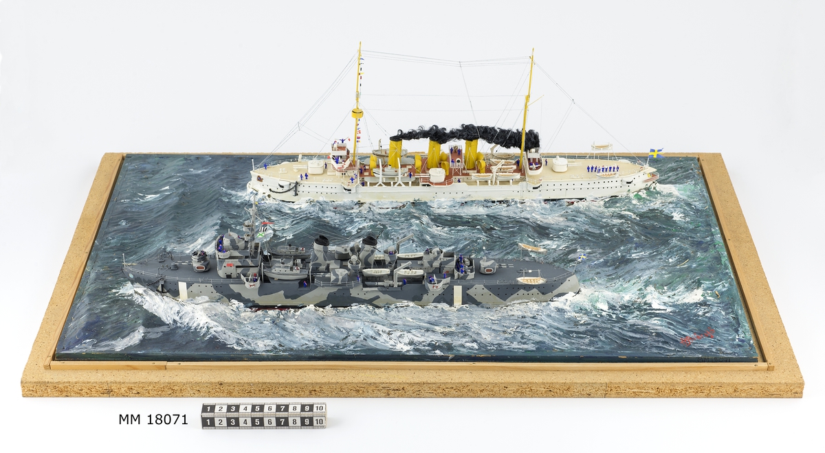 Fartygsdiorama visande två fartyg under gång till sjöss. Modellen är tillverkad av trä med detaljer av plast, papper och metall. Modellerna är placerade i ett hav av någon form av plastelina, målat i gröna och blå toner. Havet är uppbyggt på en rektangulär platta. Modellerna är detaljerat återgivna och visar pansarkryssaren "Fylgia", den ena modellen visar fartyget i sitt ursprungliga skick, den andra modellen visar fartyget efter moderniseringen 1939-40. Den "äldre" modellen visar ett vitt fartyg med rök bestående av grå ull från de tre skorstenarna, den "yngre" modellen visar ett kamouflagemålat fartyg försett med vitt neutralitetsband. Dioramat är signerat på havet med rött: "H Biärsjö".