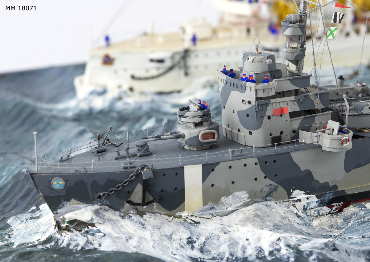 Fartygsdiorama visande två fartyg under gång till sjöss. Modellen är tillverkad av trä med detaljer av plast, papper och metall. Modellerna är placerade i ett hav av någon form av plastelina, målat i gröna och blå toner. Havet är uppbyggt på en rektangulär platta. Modellerna är detaljerat återgivna och visar pansarkryssaren "Fylgia", den ena modellen visar fartyget i sitt ursprungliga skick, den andra modellen visar fartyget efter moderniseringen 1939-40. Den "äldre" modellen visar ett vitt fartyg med rök bestående av grå ull från de tre skorstenarna, den "yngre" modellen visar ett kamouflagemålat fartyg försett med vitt neutralitetsband. Dioramat är signerat på havet med rött: "H Biärsjö".