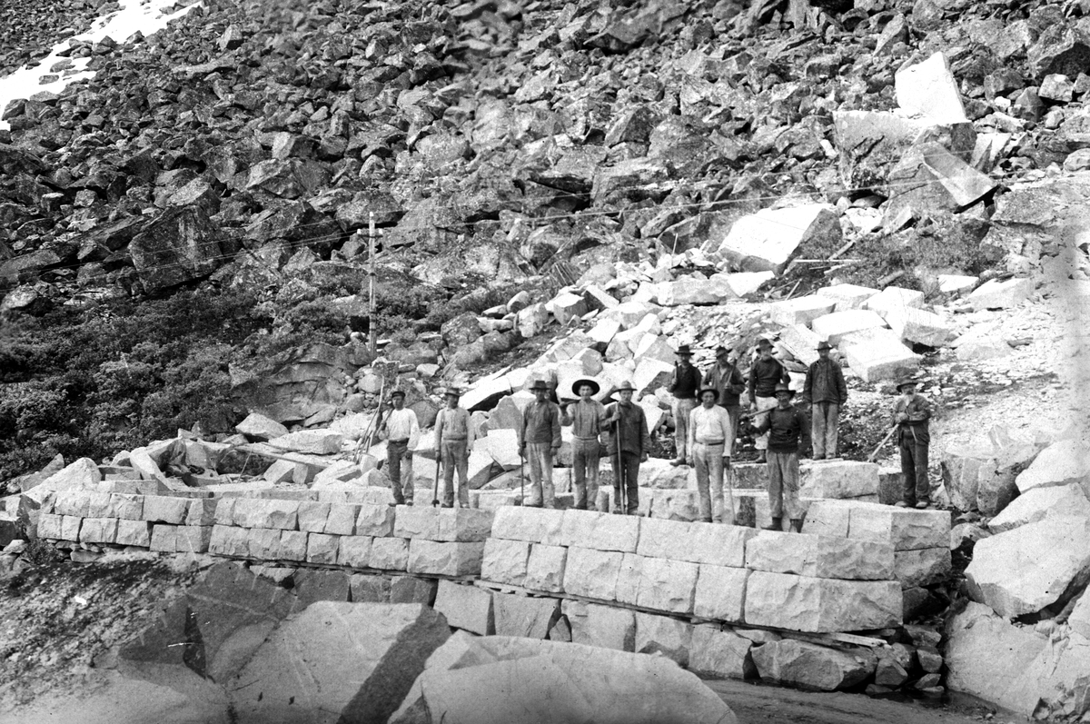 Anleggsarbeidere, steinarbeidere, steinmur, steinblokker, redskaper, ukjent sted.